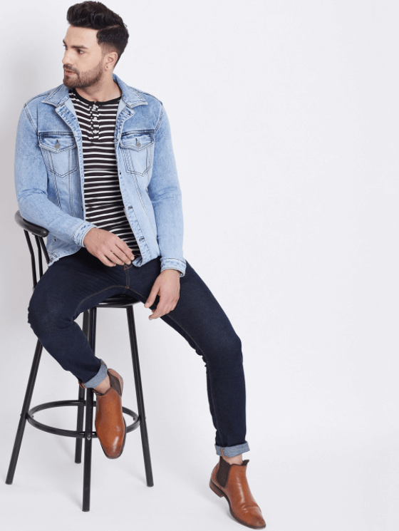 Light blue denim jacket for men with striped shirt and dark blue jeans, seated on a stool against a white background.