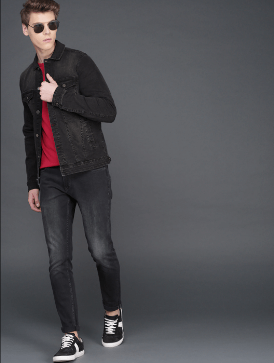 Stylish man in black denim jacket, red sweater, and sneakers posing against gray background.