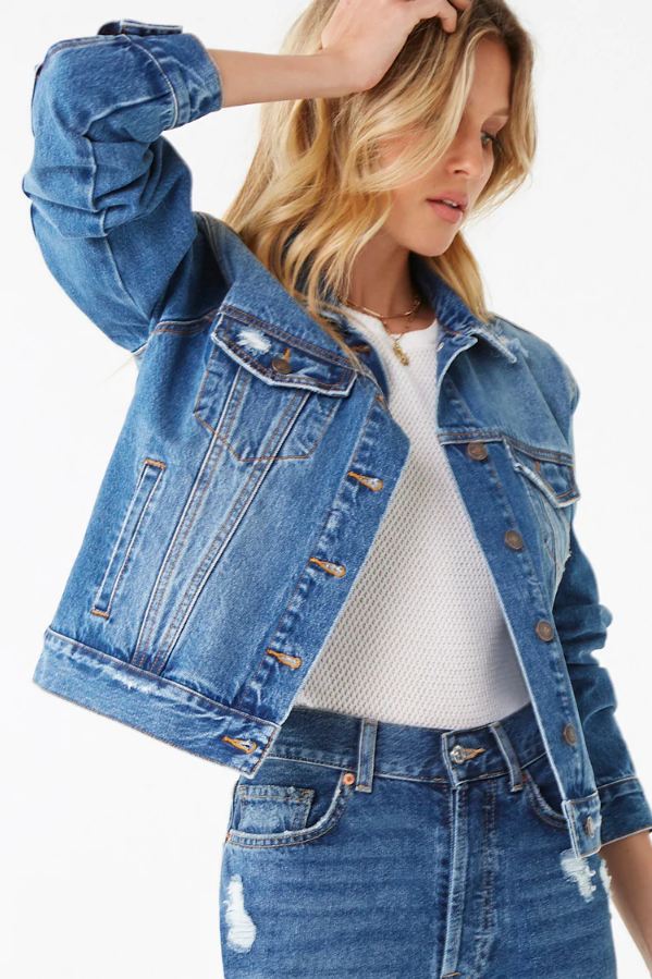 Distressed denim jacket for women, featuring a cropped silhouette and a classic blue wash. The jacket is worn over a white top, creating a stylish, casual look.
