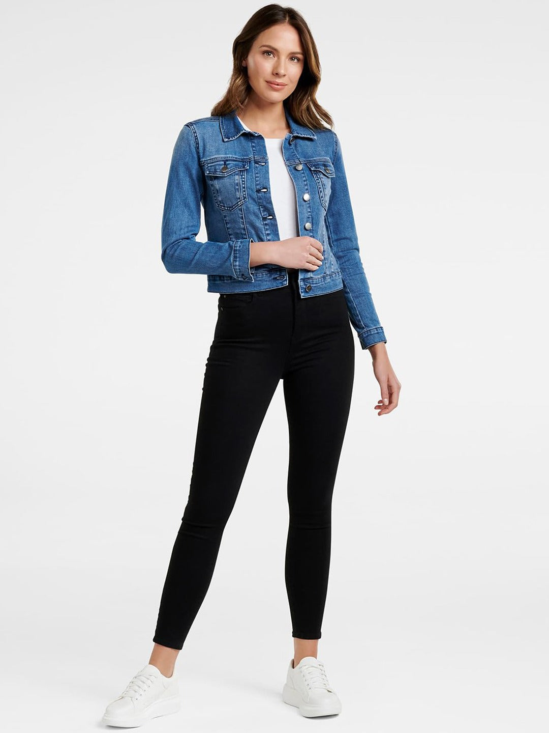 Women's blue denim jacket with front pockets, worn over black skinny jeans and paired with white sneakers.