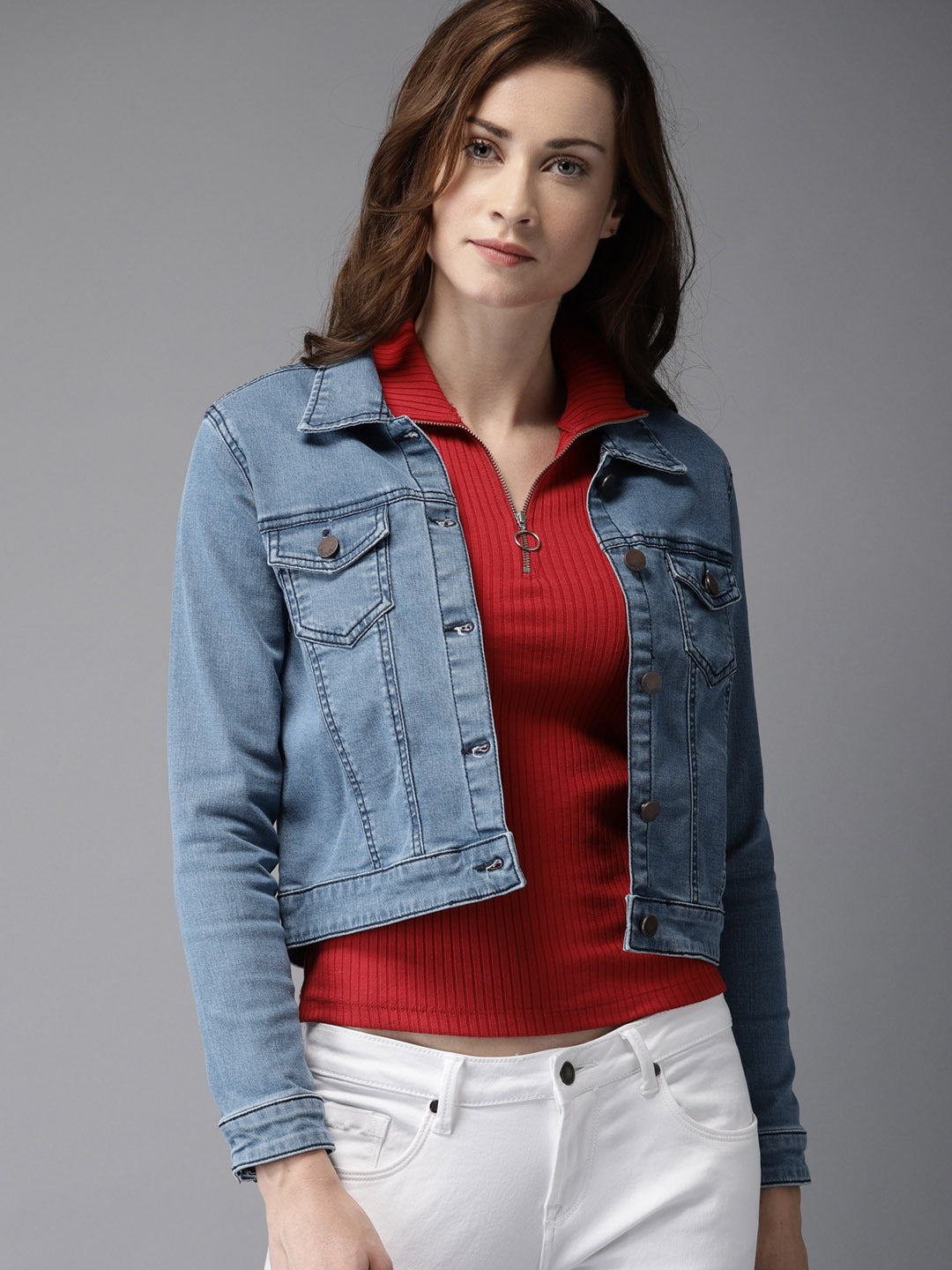 Blue denim jacket with red knit top, worn by young, brunette woman against gray background