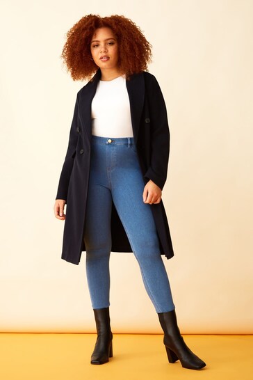 Fashionable woman in black coat, white top, and blue jeans posing confidently in a studio setting.