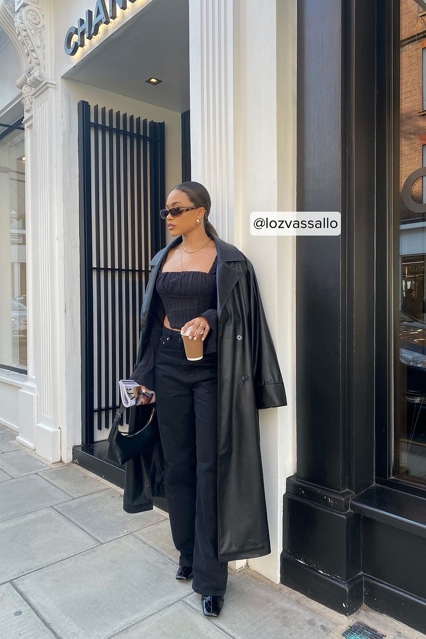 Chic black trench coat, stylish sunglasses, sleek black outfit - fashionable woman posing outside store