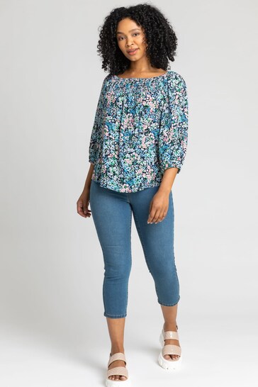 Floral blouse with puffed sleeves, blue cropped jeggings, and beige heels worn by a smiling woman with curly dark hair.