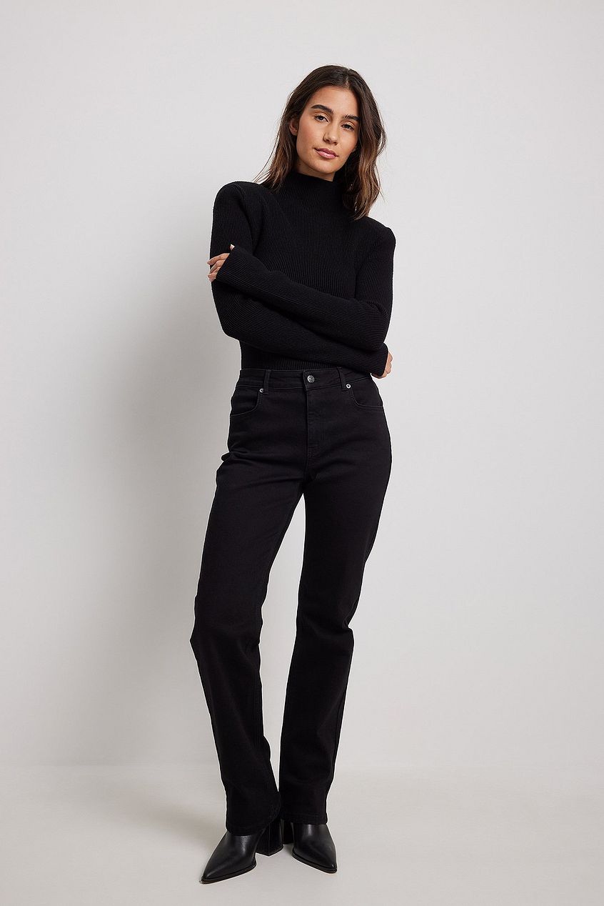 Sleek black turtleneck sweater and dark denim jeans worn by a fashion-forward woman with long brunette hair, standing against a plain white background.