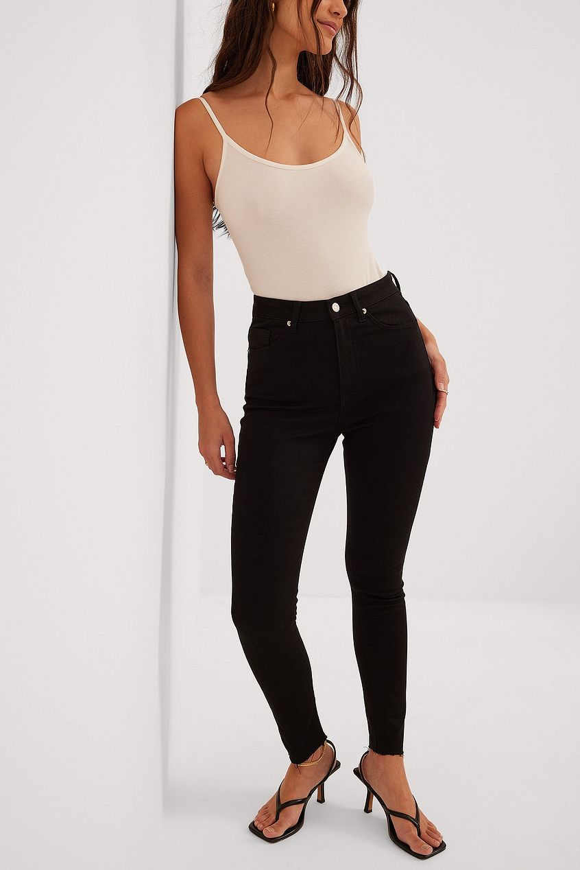 Skinny high-waist black raw-hem jeans worn by a young woman with long brown hair in a white tank top
