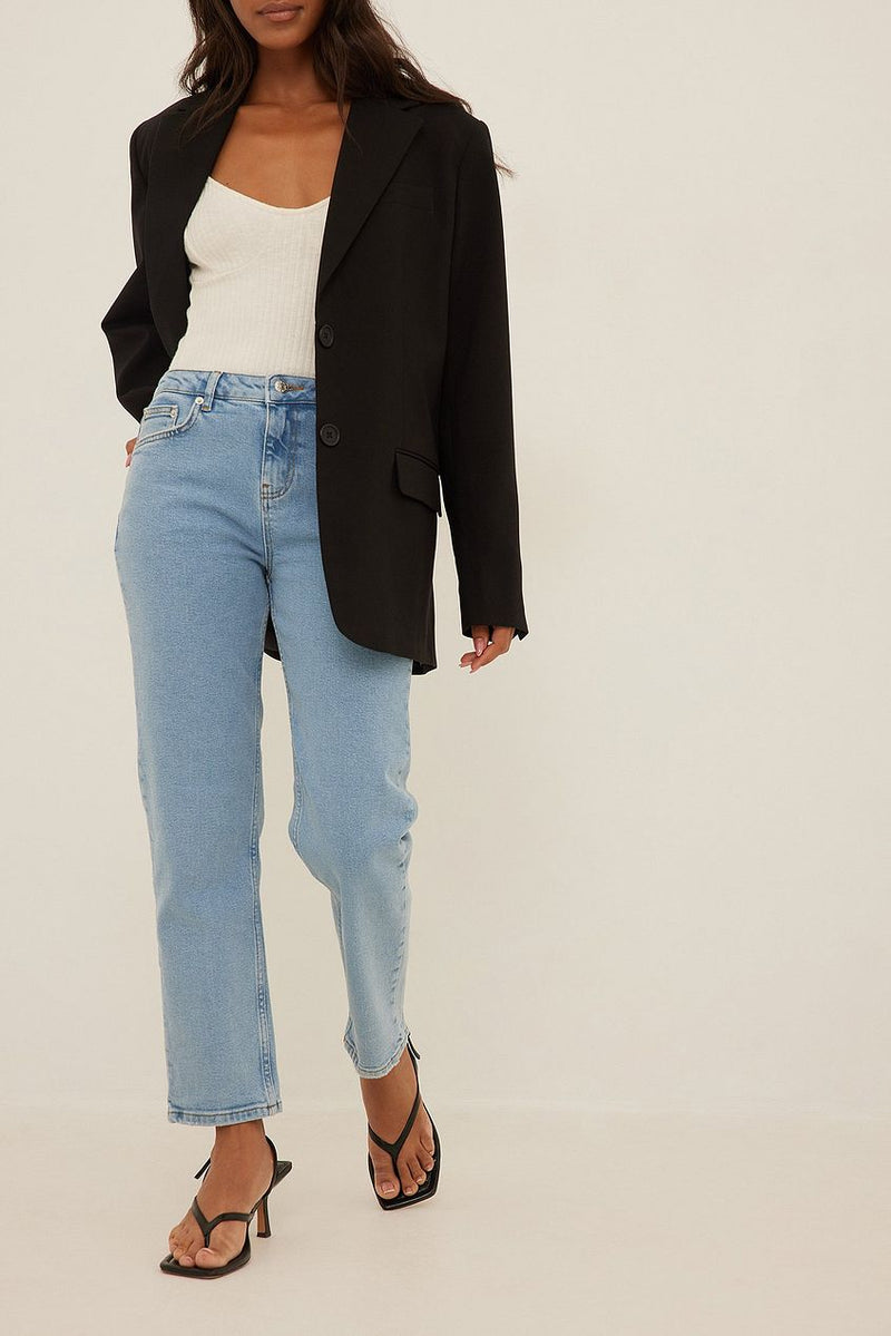 Stylish woman in mid waist slim leg jeans, white top, and black blazer posing against a white background.