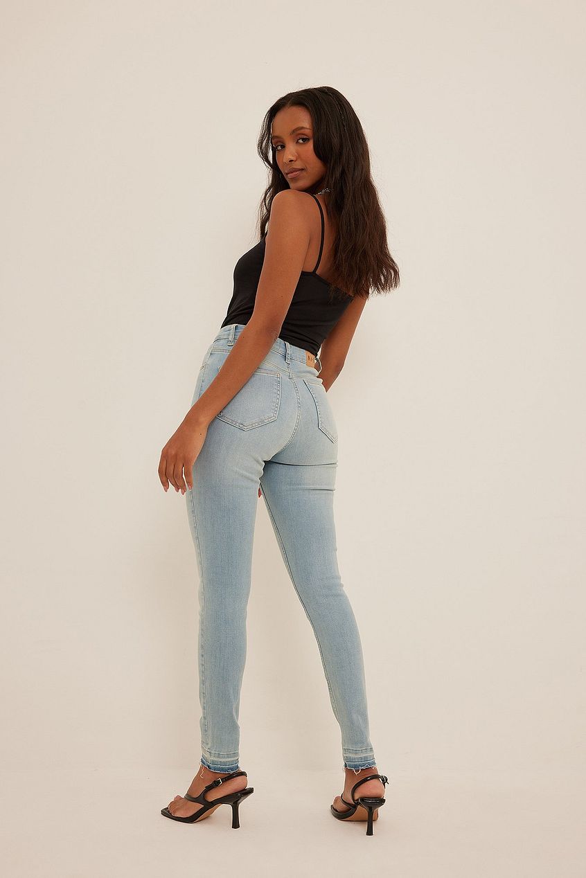 Skinny high waist open hem blue denim jeans on young woman with long dark hair posing against plain white background.