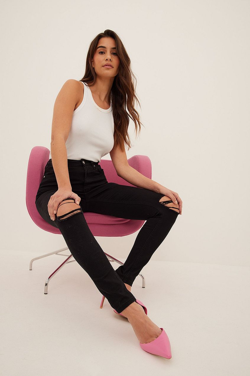 White tank top, black high-waisted destroyed skinny jeans, and pink heels - stylish and modern casual outfit worn by a young woman with long brown hair sitting on a pink chair.