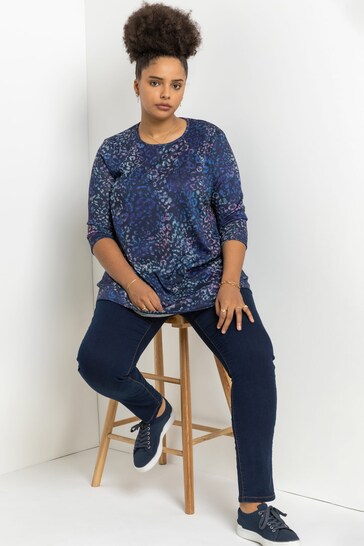 Navy blue floral print stretchy jeggings with a loose-fitting top, worn by a young woman with curly dark hair, seated on a wooden stool against a plain white background.