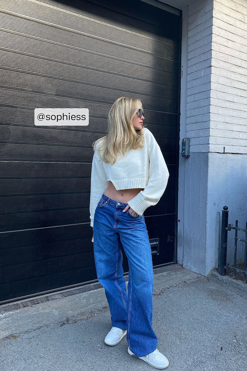 Stylish wide leg denim jeans, relaxed fit woman's blue jeans, fashionable cropped white sweater, casual street style outfit near grey garage door.