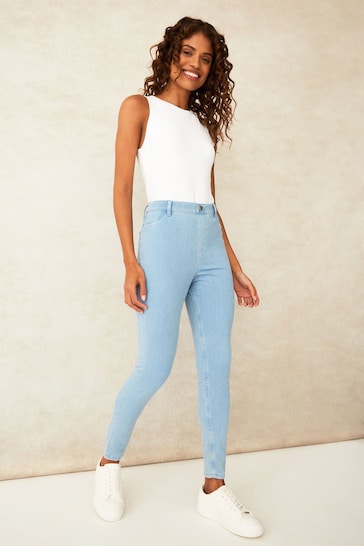 Stylish woman in white tank top and light blue skinny jeggings from Ace Cart, standing against a plain background.