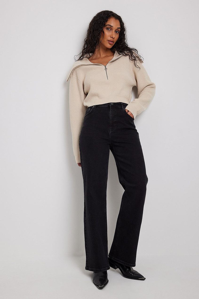 High-waist black denim jeans and beige half-zip sweater worn by young woman with long curly dark hair against white background.